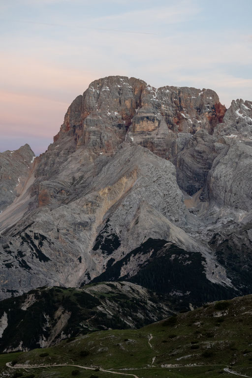 Schroffe Dolomitengipfel erheben sich über grünen Almwiesen und felsigen Hängen. Das sanfte Licht der Morgendämmerung und ein Himmel in zarten Rosa- und Blautönen verleihen der beeindruckenden Berglandschaft eine ruhige und friedliche Atmosphäre.