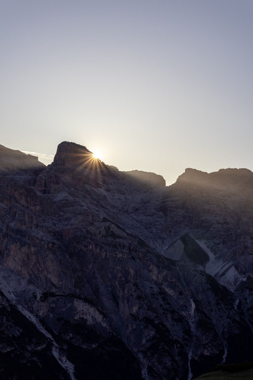 Die aufgehende Sonne schiebt sich als heller Lichtstern über einen schroffen Felsgrat der Dolomiten und taucht die Berglandschaft in sanftes Morgenlicht. Die felsigen Hänge liegen noch im Schatten und vermitteln die stille Atmosphäre eines frühen Tagesbeg
