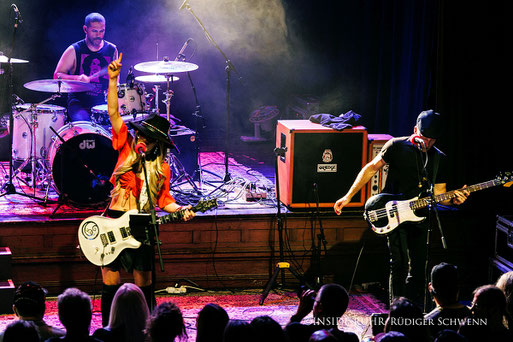 <img src="beispielbild.jpg" alt="Orianthi live im Musiktheater Piano, singt und spielt weiße E-Gitarre, flankiert von Schlagzeuger und Bassist, Publikum jubelt">