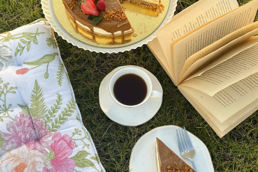 芝生の上に置かれた花柄のクッション、本、コーヒーの入ったカップ＆ソーサ、チョコレートケーキのお皿。