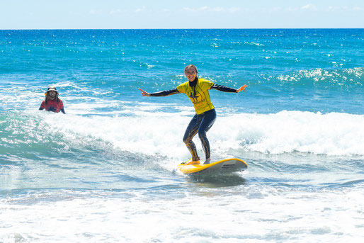 Improver surfer riding a green wave during a lesson in Gran Canaria, with instructor smiling and cheering in the background.