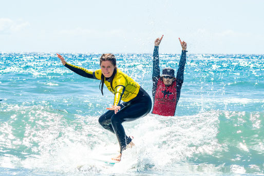 Beginner surfer riding a small wave during a surf lesson in Gran Canaria, with instructor cheering them on from the water.