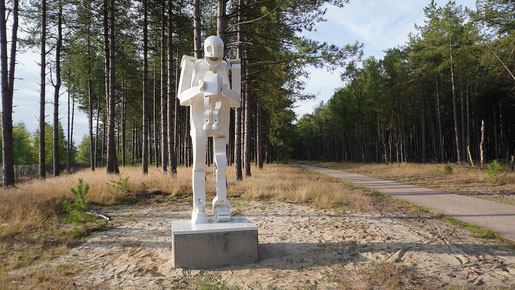 Kunst in de openbare ruimte: Saturnus: Witte sculptuur van een gestileerde figuur op sokkel in het dennenbos van de Sahara in Lommel; zandgrond en wandelpad op de achtergrond.
