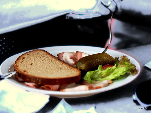 Ein Teller mit einer kleinen Bauernbrotzeit  nur Schemenhaft ein Blauer Hintergrund, warm leuchtet das Bauernbrot und der Räucherschinken mit Gurke. 