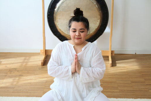 Frau in Weiß sitzend und meditierend. Ein Gong im Hintergrund.