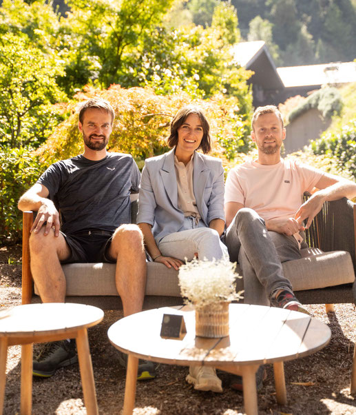Drei lächelnde Personen sitzen gemeinsam auf einer Outdoor-Lounge in sonniger Gartenlandschaft, umgeben von üppigem Grün und kleinen Holztischen.