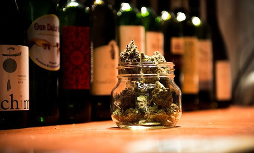 Ein Glas voll mit Cannabis Blüten steht auf einer Holztheke. Im Hintergrund stehen viele Flaschen Wein.