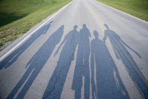 Sechs Personen laufen auf einer leeren Straße. Sie sind nur als Schatten auf dem Asphalt zu sehen. Obwohl man nur die Umrisse erkennt, wirkt die Gruppe fröhlich und vertraut.