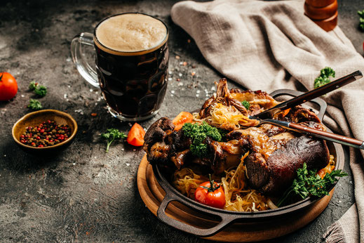 Gußpfanne mit zubereitetem Eisbein auf Sauerkraut dekoriert mit Kräutern und Strauchtomate, im Hintergrund eine kleine Schale mit bunten Pfefferkörnern und ein Glas Dunkelbier