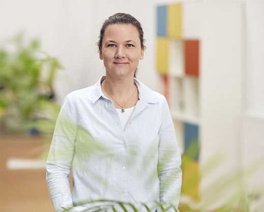 Farbportrait von Ines Walter. Sie trägt ein blau-weiß fein-gestreiftes Hemd und eine Kette mit grünem Stein. Sie trägt ihre Haare zurückgebunden und lächelt in die Kamera. Im Hintergrund ist eine grüne unscharfe Pflanze und ein buntes Regal zu sehen.