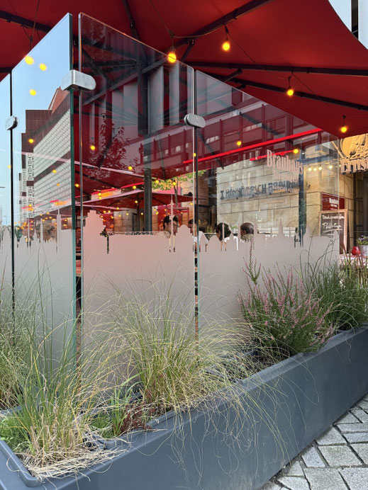 Glas-Windschutz mit individueller Logo-Folierung im Skyline-Design und integrierten Blumenkästen für die Außengastronomie