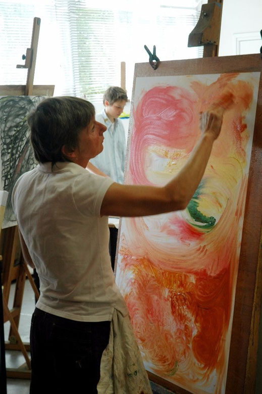 een vrouw staat voor een groot schilderij op papier, voor een ezel te werken. Ze schildert vooral met rode en gele tinten. Ze schildert met haar handen in de verf.