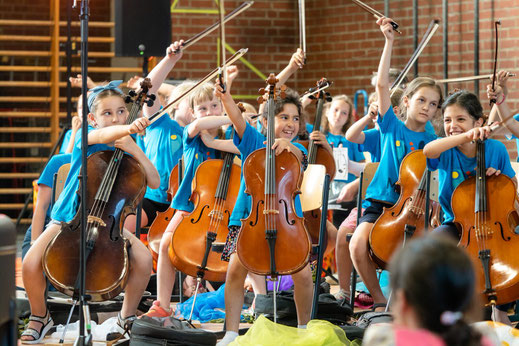 ca. 10 Cellist*innen des Musaik-Orchesters heben freudig ihre Cellobögen in die Luft.