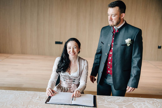 Hochzeit Sistrans Standesamt Trauung Braut sitzt am Tisch unterschreibt Urkunde Bräutigam steht hinter ihr und schaut Braut an