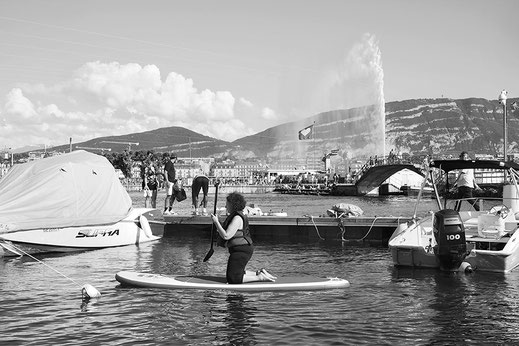 Photographie noir et blanc. Genève, vue du bains des Pâquis sur le Léman, le jet d'eau, le Salève, à l'avant-plan une dame à genoux sur un paddle. 