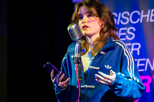 Poetin Mali Carillo steht mit ihrem Handy in der Hand am Mikrofon und präsentiert ihren Text.