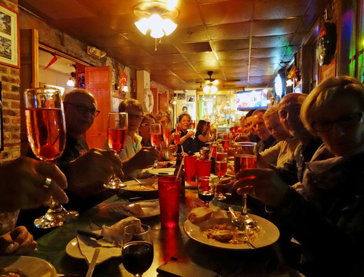 Motoglobe_Motorradreisen. Viele Leute sitzen an einem Tisch im Restaurant und haben Essensteller vor sich auf dem Tisch und prosten sich alle mit einem Glas zu.