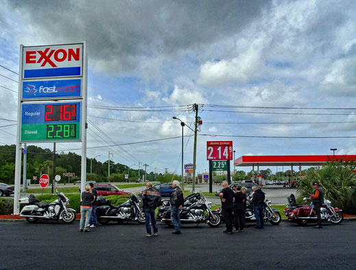 Motoglobe_Motorradreisen. Die Motorräder stehen zwischen zwei grossen Tankstellen auf einem Teerplatz und die Fahrer stehen dazwischen und reden miteinander.
