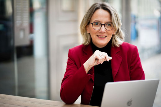Foto: Sabine Lansing, Knigge-Expertin sitzt an einem Tisch, vor sich ein Apple MacBook
