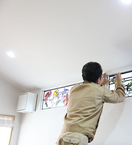 ステンドグラス専門技術者が住宅の高窓にステンドグラスパネルを取り付けている様子。明るい室内で繊細なガラス作品を丁寧に施工している風景。