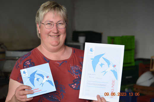 Frau Ermann hält in ihrer einen Hand (vom Leser aus links) die Plakette für "Schwalbenfreundliches haus". In ihrer anderen Hand hält sie die dazugehörige Urkunde.