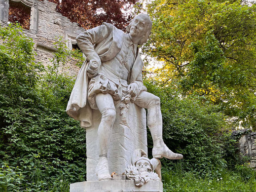 Photograph of a stone monument to William Shakespeare in a seated pose in Ilmpark, surrounded by trees