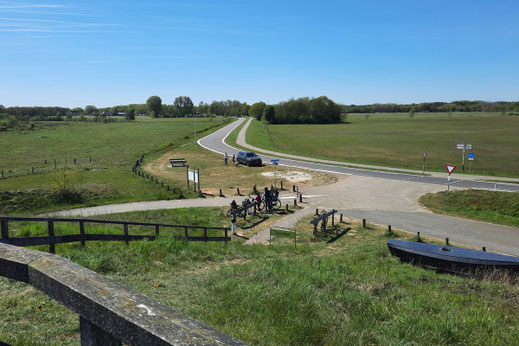 Eine Straße führt durch eine Ebene. Neben ihr ist ein breiter Fahrradweg angelegt. In der Kurve ist ein Rastplatz angelegt. 