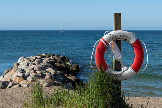 An einem Strand hängt ein Rettungsring an einem Holzpfahl. Im Hintergrund ist das blaue Meer mit einem Seegelboot zu sehen.