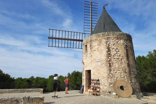 Eine Frau steht vor eine alten Windmühle auf einem Hügel. Neben dem Eingang zur Mühle steht ein Ständer mit Andenken.
