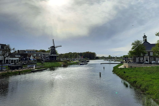 An einem Fluss stehen im Abendlicht eine alte Windmühle und ein paar Häuser.