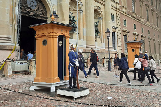 Soldaten in blauen Gardeuniformen stehen vor Wachhäuschen vor einem Schloss. Passanten gehen in das Schloss hinein.stehen vor
