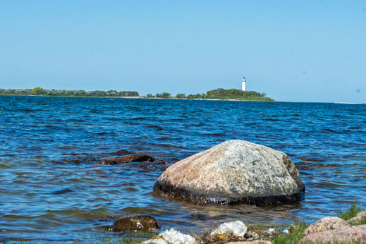 Im Meer liegt ein großer Fels. Am anderen Ende der Bucht steht, etwas hinter einem Wald verborgen, ein Leuchtturm.