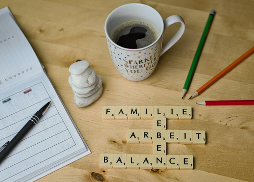 Ein Kaffeebecher steht auf einen Tisch, davor wurden mit Scrabble die Worte Familie, Arbeit, Balance und Leben gelegt