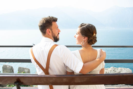 Das Brautpaar sitzt auf einer Bank und dieht sich an. Im Hintergrundf der Gardasee in Italien.