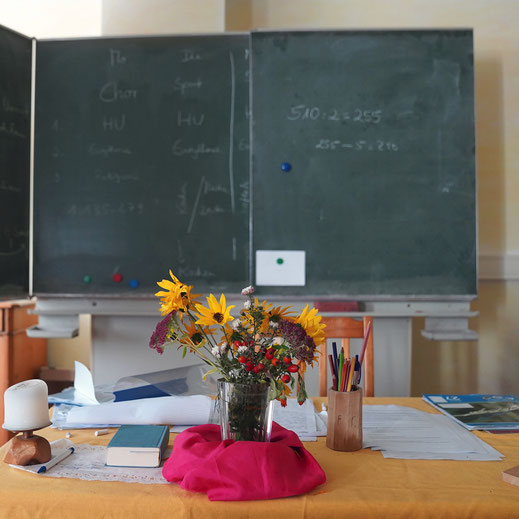 Ein Foto auf einen Tisch im Klassenzimmer auf dem Blumen stehen und ein Buch liegt. Im HIntergrund sieht man eine Schultafel.