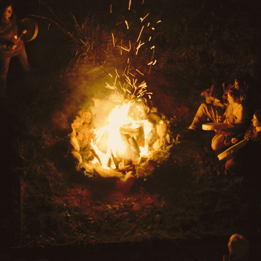 Menschen sitzen nachts im Kreis um ein loderndes Lagerfeuer, Funken steigen in die Dunkelheit