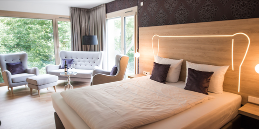 Modern eingerichtetes Hotelzimmer mit großem Doppelbett, eleganter Sitzecke und großen Fenstern mit Blick ins Grüne.