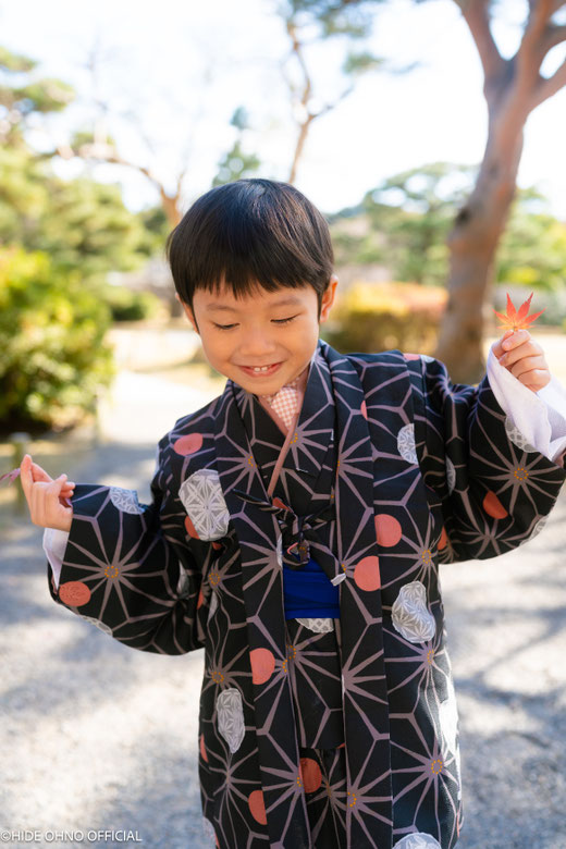Smiling boy in black kimono holding a red maple leaf — charming autumn photoshoot in Tokyo.