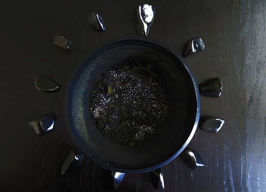 Eine schwarze Schungit Schüssel gefüllt mit einem schwarzen Pulver und silbernem Glitzer umgeben von Schungit Steinen, auf schwarzem Holz