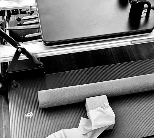 A black and white photo of STUDIO ORIGIN Pilates’ interior, tilted to the floor to show examples of high quality equipment like a Pro reformer next to a Professional deeply cushioned mat, cushioning soft foam roller, cork blocks and stretch band.