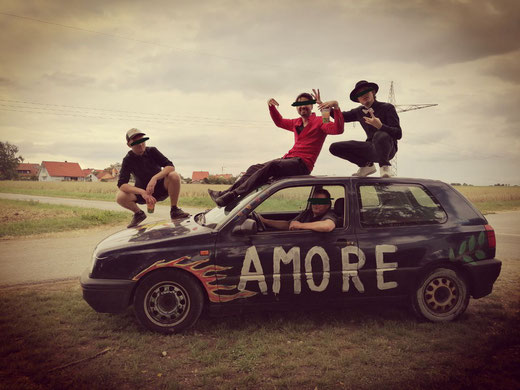 Die vier Bandmitglieder posieren auf und in einem Auto, auf dem Amore steht.