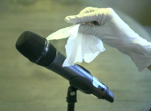 Handheld wireless microphone in a stand with woman's hand wearing a rubber glove holding a sanitizing wipe near the microphone.