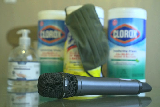 Wireless handheld microphone on a table with a pump hand sanitizer dispenser and three containers of sanitizing wipes in the background.