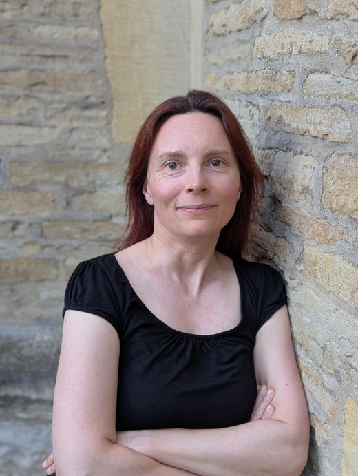 Mittelalter Frau mit braunen Haaren, die sich an eine Mauer lehnt und freundlich lächelt.
