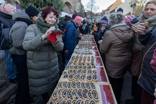 Im Freien steht ein Tisch, auf dem gleichmäßig geschnittene Scheiben Mohnstollen bereitstehen. Um den Tisch herum hat sich das Publikum der Mohnstollenverkostung versammelt und betrachtet und kostet den Mohnstollen.