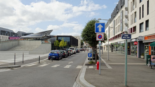 La rue Nicolas Bouvier, située au Nord de la Médiathèque. Sur la gauche de la photo, la piste cyclable qui lone la médiathèque. Sur la droite, le large trottoir qui longe les commerces