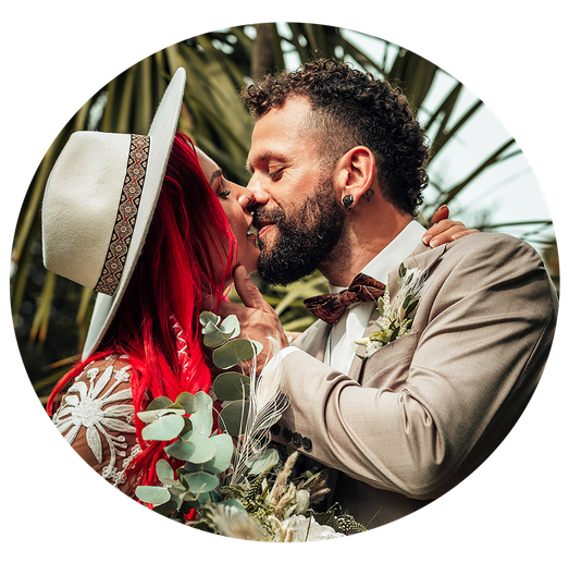 Boho Braut mit rotem Haar küsst ihren bärtigen Bräutigam unter Palmen. Ihr Hut, sein beiger Anzug & der Eukalyptusstrauß - pure, wilde Hochzeitsromantik