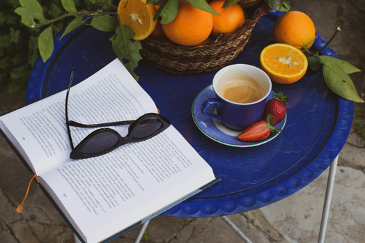 青色のガーデンテーブルに広げられた本とサングラス。オレンジが盛り付けられた籐のかご。いちごがソーサーに添えられたコーヒーカップ。