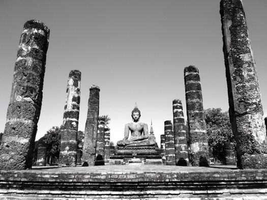 Das Bild ist schwarz-weiß. Es ist eine Buddhastatue auf einer Steinruine zu sehen. Links und rechts von ihm stehen jeweils fünf Steinsäulen.
