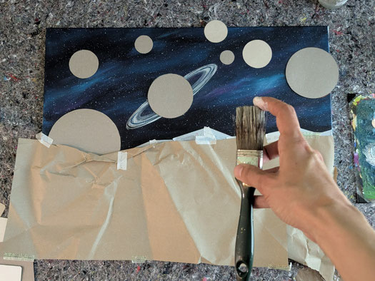 Acrylmalerei-Technik: Hand hält Pinsel und spritzt Sterne auf einen dunkel gestalteten Himmel mit Planeten. 